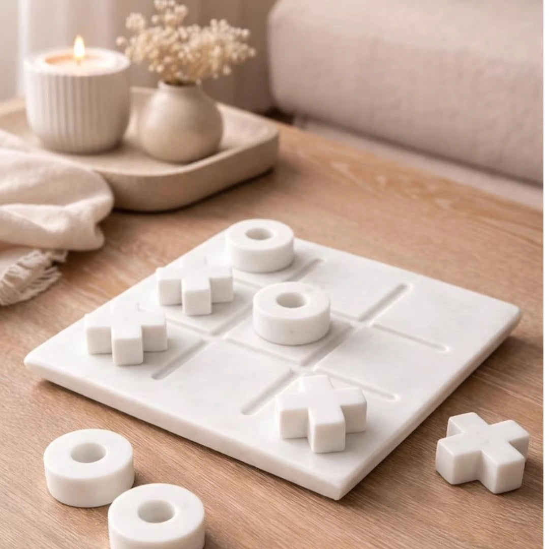 White marble tic tac toe game set with smooth X and O pieces arranged on a square board, styled on a neutral beige surface with soft fabric and floral decor.