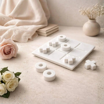 White marble tic tac toe game set with smooth X and O pieces arranged on a square board, styled on a neutral beige surface with soft fabric and floral decor.